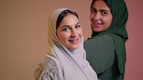 مفهوم الاناقة والاهتمام بالمظهرالخارجي، الشغف باتباع صيحات الموضة، عارضة عربية خليجيه سعودية محجبة ترتدي عباءه انيقة عصرية تقف برفقه صديقتها، الوقوف بالجانب والنظرالى الكاميرا بتعابير الثقة بالنفس، امرأه اماراتيه مبتسمه تقوم برتيب الحجاب