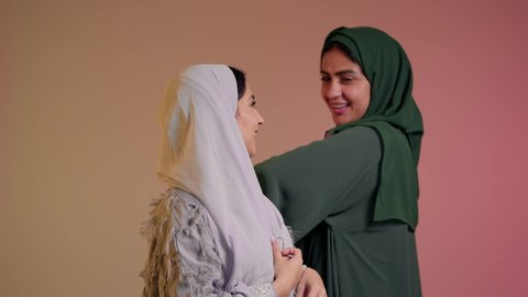 مفهوم الاناقة والاهتمام بالمظهرالخارجي، الشغف باتباع صيحات الموضة، عارضة عربية خليجيه سعودية محجبة ترتدي عباءه انيقة عصرية تقف برفقه صديقتها، الوقوف بالجانب والنظرالى الكاميرا بتعابير الثقة بالنفس، امرأه اماراتيه مبتسمه تقوم برتيب الحجاب