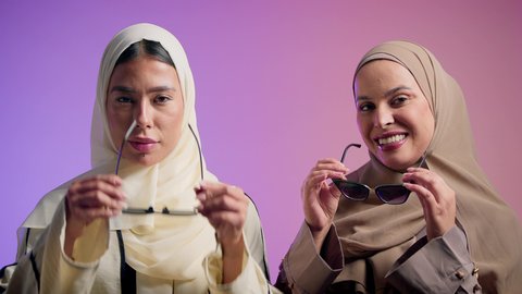 أزياء محجبات عصرية راقية، مفهوم الاهتمام بالأناقة والمظهر الخارجي، بورترية مقرب لصديقتان عربيتان خليجيتان اماراتيان تقومان بوضع النظارة الشمسية، سيدتان سعوديتان تنظران الى الكاميرا بايماءات الثقه و الاعتزاز بالنفس ، خلفية ملونه