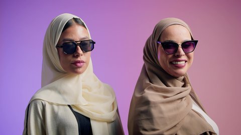 Gestures indicating femininity and beauty, a close-up portrait of two Arab Gulf Emirati friends wearing colorful abayas and sunglasses, two Saudi women looking at the camera with gestures of confidence and self-esteem, colorful background