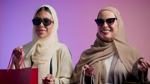 Benefit from the Black Friday offers and discounts, fashion, elegance and interest in the external appearance, a close-up portrait of two smiling veiled Saudi Arabian Gulf girls wearing colorful abaya and sunglasses looking at the camera with gestures of pleasure, two Saudi friends holding colorful shopping bags in their hands, colorful background