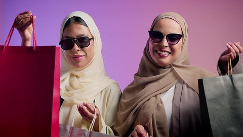 الاستمتاع بالشراء والتبضع، الموضة والأناقة والاهتمام بالمظهر الخارجي، بورترية مقرب لشابتان عربيتان خليجتان سعوديتان محجبتان مبتسمتان ترتديان العباءة الملونه و النظارة الشمسية تنظران إلى الكاميرا بإيماءات السرور، صديقتان سعوديتان تحملان بيديهما اكياس التسوق ملونه ، خلفية ملونه