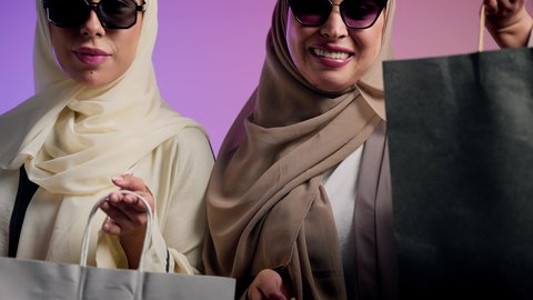متعة التسوق في المولات، الموضة والأناقة والاهتمام بالمظهر الخارجي، بورترية مقرب لشابتان عربيتان خليجتان سعوديتان محجبتان مبتسمتان ترتديان العباءة الملونه و النظارة الشمسية تنظران إلى الكاميرا بإيماءات السرور، صديقتان سعوديتان تحملان بيديهما اكياس التسوق ملونه ، خلفية ملونه