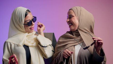 Seasonal offers and discounts in the markets, the concept of softness, chic and femininity, fashion, elegance and interest in external appearance, a close-up portrait of two smiling veiled Saudi Arabian Gulf girls wearing colorful abayas, looking at the camera with gestures of pleasure and self-confidence, two Saudi friends holding colorful shopping bags in their hands, colorful background