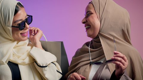 العروض والتخفيضات السنوية في الأسواق، مفهوم النعومة والشياكة والأنوثة، الموضة والأناقة والاهتمام بالمظهر الخارجي، بورترية مقرب لشابتان عربيتان خليجتان سعوديتان محجبتان مبتسمتان ترتديان العباءة الملونه تنظران إلى الكاميرا بإيماءات السرور والثقة بالنفس،سيده سعودية تحمل بيديها اكياس تسوق ملونه ، خلفية ملونه