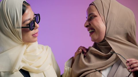 عباءات خليجية نسائية عصرية، مفهوم النعومة والشياكة والأنوثة، الموضة والأناقة والاهتمام بالمظهر الخارجي، بورترية مقرب لشابتان عربيتان خليجتان سعوديتان محجبتان مبتسمتان ترتديان العباءة الملونه تنظران إلى الكاميرا بإيماءات السرور والثقة بالنفس، صديقتان اماراتيان يقومان بقضاء أوقات ممتعة ، خلفية ملونه