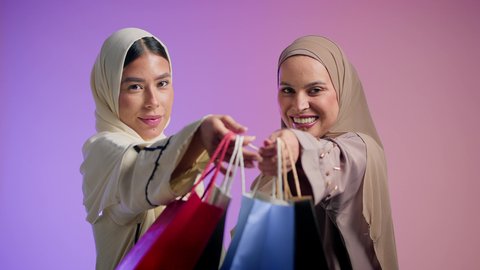 Taking advantage of discounts and exciting offers, Black Friday offers, buying gifts and personal supplies, close-up portrait of two Saudi Arabian Gulf women wearing colorful hijab and abaya carrying shopping bags, two Emirati friends enjoying shopping in malls and shops, colorful background