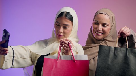 مفهوم التسوق برفقة الأصدقاء، قضاء أوقات ممتعة في شراء الهدايا والمستلزمات، بورتريه لشابتين عربيتين خليجيتين سعوديتين محجبتين ترتديان العباءات العصرية تمسكان بالأكياس الورقية، امراة امارتية تقوم بالتقاط صور سيلفي باستخدام الهاتف المحمول، خلفية ملونة
