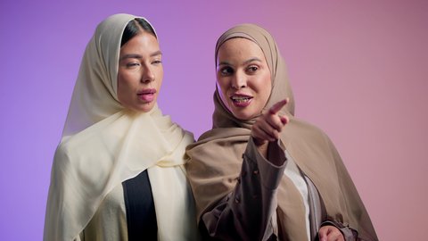 الوقوف باستقامة والنظر إلى شيء ما، الدردشة والتسامر مع الأصدقاء، سيده عربيه خليجيه اماراتية تقوم بالاشارة الى شيء ما برفقه صديقتها ، بورتريه مقرب لشابتان سعوديتان محجبتان يقومان بقضاء اوقات ممتعه ، ايماءات السعادة و السرور، خلفية ملونة