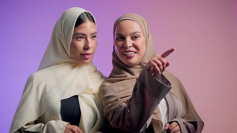 تحديد موقع شيء معين، الدردشة والتسامر مع الأصدقاء، سيده عربيه خليجيه اماراتية تقوم بالاشارة الى شيء ما برفقه صديقتها ، بورتريه مقرب لشابتان سعوديتان محجبتان يقومان بقضاء اوقات ممتعه ، ايماءات السعادة و السرور، خلفية ملونة