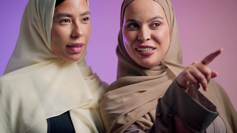 التوضيح والدلالة لمكان محدد، الدردشة والتسامر مع الأصدقاء، سيده عربيه خليجيه اماراتية تقوم بالاشارة الى شيء ما برفقه صديقتها ، بورتريه مقرب لشابتان سعوديتان محجبتان يقومان بقضاء اوقات ممتعه ، ايماءات السعادة و السرور، خلفية ملونة