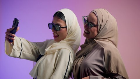 استخدام التقنيات الحديثة، الإمساك بالحقائب الجلدية الفاخرة ذات البراندات العالمية، سيدة عربيه خليجيه اماراتية تقوم التقاط صور السيلفي برفقة صديقتها ، بورتريه مقرب لشابتان سعوديتان محجبتان ترتديان العباءات الملونة و النظارات الشمسية تقفان وتأخذان اللقطات باستخدام الهاتف المحمول وصنع ذكريات جميلة، خلفية ملونة