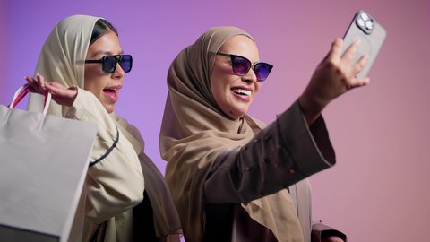 الإمساك بالحقائب الجلدية الفاخرة ذات البراندات العالمية، سيدة عربيه خليجيه اماراتية تقوم التقاط صور السيلفي برفقة صديقتها ، بورتريه مقرب لشابتين سعوديتين محجبتين ترتديان العباءات الملونة و النظارات الشمسية تقفان وتأخذان اللقطات باستخدام الهاتف المحمول وصنع ذكريات جميلة، خلفية ملونة