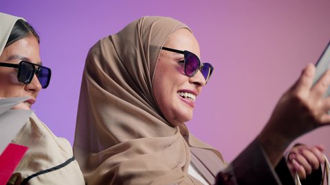 الاستمتاع بالشراء والتسوق ، سيدة عربيه خليجيه اماراتية تقوم التقاط صور السيلفي برفقة صديقتها ، بورتريه مقرب لشابتين سعوديتين محجبتين ترتديان العباءات الملونة و النظارات الشمسية  تقفان وتأخذان اللقطات باستخدام الهاتف المحمول وصنع ذكريات جميلة، الاهتمام بالأناقة والمظهر الخارجي، خلفية ملونة