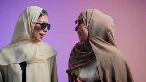 الاعتزاز و الثقة بالنفس، أزياء محجبات عصرية أنيقة، الاهتمام بالمظهر الخارجي و الأناقة، بورترية مقرب لإمرأة عربية خليجية اماراتية ترتدي الحجاب والعباءة الملونه برفقه صديقتها تنظر الى الكاميرا بإيماءات السعادة ، شابتان سعوديتان ترتديان النظارات الشمسية تقضيان أوقات ممتعة ومسلية، خلفية ملونه