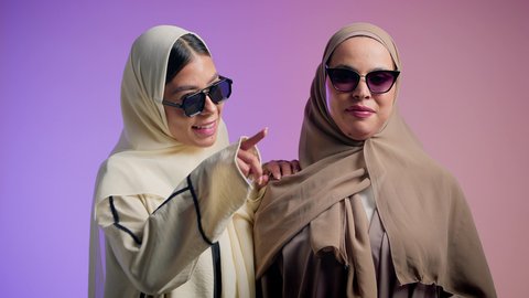 مفهوم الأناقة والشياكة و الأنوثة والنعومة، بورترية مقرب لإمرأة عربية خليجية اماراتية ترتدي الحجاب والعباءة الملونه  تتحدث مع صديقتها و تشاور الى شيء ما بإيماءات السعادة، شابتان سعوديتان  ترتديان النظارات الشمسية تقضيان أوقات ممتعة ومسلية، خلفية ملونه