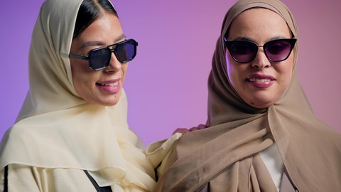 المودة و المحبة بين الأصدقاء، بورترية مقرب لإمرأة عربية خليجية اماراتية ترتدي الحجاب والعباءة الملونه  تتحدث مع صديقتها و تشاور الى شيء ما بإيماءات السعادة، شابتان سعوديتان  ترتديان النظارات الشمسية تقضيان أوقات ممتعة ومسلية، خلفية ملونه