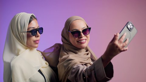 الاهتمام بالأناقة والمظهر الخارجي،صديقتان سعوديتان تقومان بالتقاط صور السيلفي برفقة الأصدقاء وقضاء أوقات ممتعة، بورتريه لشابتان عربيتان خليجيتان اماراتيان محجبتان ترتديان العباءات الملونة و النظارات الشمسية تقفان وتأخذان اللقطات باستخدام الهاتف المحمول وصنع ذكريات جميلة، خلفية ملونة