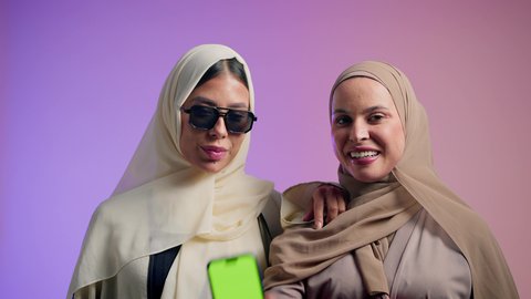 Chroma key, use of modern technical devices, interest in elegance and outward appearance, close-up portrait of a smiling veiled Saudi Arabian Gulf woman wearing a colorful abaya displaying a blank green screen via mobile phone with her friend, an Emirati Gulf woman wearing sunglasses standing with confidence and pride, colorful background