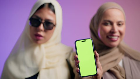 استخدام الأجهزة التقنية الحديثة، الاهتمام بالأناقة والمظهر الخارجي، بورتريه مقرب لامرأة عربية خليجية سعودية مبتسمة محجبة ترتدي العباءة الملونه تقوم بعرض شاشة خضراء فارغة عبر الجوال برفقه صديقتها, سيده خليجيه اماراتية ترتدي النظارة الشمسيه تقف بثقه و اعتزاز, خلفية ملونه