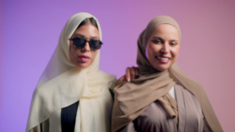 Mobile phone screen display, portrait of two Arab Gulf Emirati friends wearing colorful abaya and sunglasses standing with confidence and pride, Saudi woman holding mobile phone in her hand, looking at the camera with gestures of pleasure and happiness, interest in outward appearance, colorful background