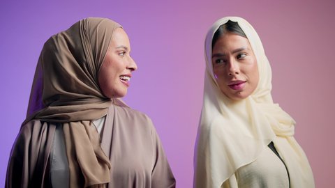 Softness, elegance and femininity, the concept of fashion, elegance and interest in outward appearance, a close-up portrait of two veiled, smiling Emirati Gulf Arab girls wearing colorful abayas, looking at the camera with gestures of pleasure and self-confidence, modern Gulf women's abayas, colorful background