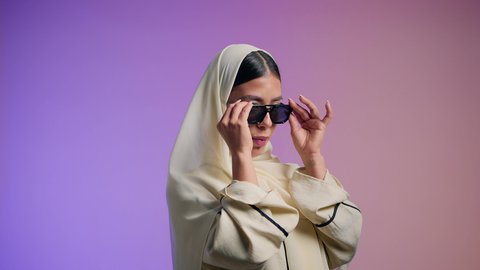 مفهوم العناية بالجمال والإطلالة المتميزة، الاهتمام بالأناقة والمظهر الخارجي، بورترية لسيدة عربية خليجية اماراتية محجبة تقوم بوضع النضارة الشمسية، سيدة سعودية تقوم بالتزين بالحلي والاكسسوارات، خبيرة مستحضرات تجميلية، خلفية ملونه