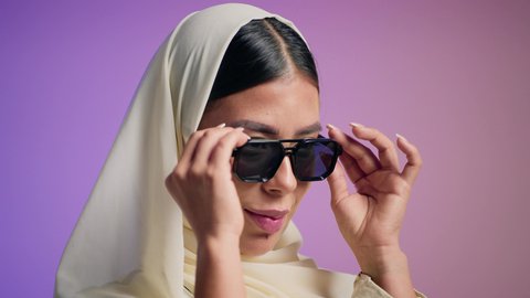 تصوير لإمرأة عربية خليجية سعودية محجبة تقوم بتجربة النظارات الشمسية الجديدة، الاهتمام بالمظهر الخارجي ، إيماءات الانوثة والجمال, الاستمتاع بإمتلاك اكسسوارات عصرية, مفهوم الموضة والاناقة ، سيدة اماراتية ترتدي عباءة أنيقة ونظارة سوداء،  خلفية ملونة