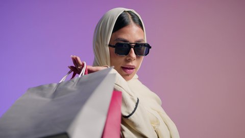 تصوير لسيده عربية خليجية سعودية مبتسمة ترتدي العباءه والحجاب تحمل بيدها أكياس التسوق الملونه، شراء المستلزمات والتبضع من المولات التجارية، سيدة اماراتية تستفيد من العروض الموسمية والتخفيضات،  الأثر الإيجابي للتسوق على السيدات، خلفية ملونه