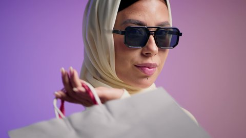 شراء المستلزمات والتبضع من المولات التجارية، سيدة اماراتية تستفيد من العروض الموسمية والتخفيضات، تصوير لسيده عربية خليجية سعودية مبتسمة ترتدي العباءه والحجاب تحمل بيدها أكياس التسوق الملونه،  الأثر الإيجابي للتسوق على السيدات، خلفية ملونه