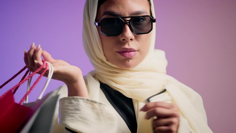 الأثر الإيجابي للتسوق على السيدات، شراء المستلزمات والتبضع من المولات التجارية، سيدة اماراتية تستفيد من العروض الموسمية والتخفيضات، تصوير لسيده عربية خليجية سعودية مبتسمة ترتدي العباءه والحجاب تحمل بيدها أكياس التسوق الملونه، خلفية ملونه