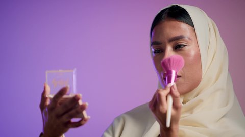 The concept of femininity and interest in appearance. Makeup and makeup tutorial. Marketing of beauty products. Photograph of a Saudi Arabian Gulf girl wearing an abaya and hijab holding a blush brush. Emirati woman using modern women's cosmetics. Elegant women's salon. Colorful background.