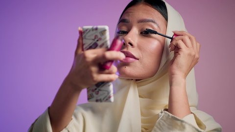 Emirati woman wearing an elegant abaya and using cosmetics. International women's products. The concept of elegance and interest in appearance. Eyeliner using mascara. Photograph of a veiled Saudi Arabian Gulf woman enjoying feminine makeup. Colorful background.