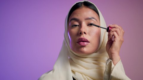 Eye drawing with mascara. International women's products. The concept of elegance and interest in appearance. A photograph of a veiled Saudi Arabian Gulf woman enjoying feminine grooming. An Emirati woman wearing an elegant abaya and using cosmetics. A colorful background.