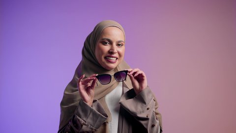 Femininity and beauty gestures: Emirati woman wearing an elegant abaya and contemporary black sunglasses. Photograph of a veiled Saudi Arabian Gulf woman trying on new sunglasses, showing interest in appearance and elegance. Colorful background.