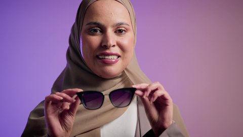سيدة اماراتية ترتدي عباءة أنيقة ونظارة سوداء معاصرة، تصوير لإمرأة عربية خليجية سعودية محجبة تقوم بتجربة النظارات الشمسية الجديدة، الاستمتاع بإمتلاك اكسسوارات عصرية,  الاهتمام بالمظهر الخارجي والاناقه ، إيماءات الانوثة والجمال,  خلفية ملونة