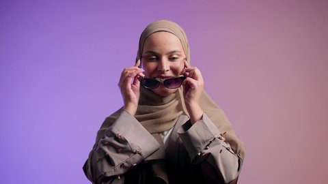 الوقوف باستقامه وثقه بالنفس ، الاستمتاع بإمتلاك اكسسوارات عصرية,  الاهتمام بالمظهر الخارجي والاناقه ،سيدة اماراتية ترتدي عباءة أنيقة ونظارة سوداء معاصرة, إيماءات الانوثة والجمال, تصوير لإمرأة عربية خليجية سعودية محجبة تقوم بتجربة النظارات الشمسية الجديدة,  خلفية ملونة