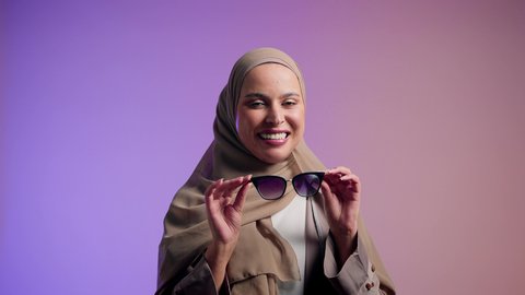 الاستمتاع بإمتلاك اكسسوارات عصرية,  الاهتمام بالمظهر الخارجي والاناقه ،تعابير السرور والسعادة ، سيدة اماراتية ترتدي عباءة أنيقة ونظارة سوداء معاصرة, إيماءات الانوثة والجمال, تصوير لإمرأة عربية خليجية سعودية محجبة تقوم بتجربة النظارات الشمسية الجديدة,  خلفية ملونة