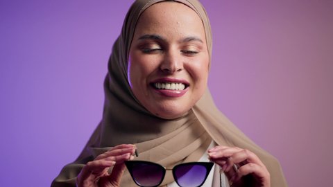 الاهتمام بالمظهر الخارجي والاناقه ،تعابير السرور والسعادة ، سيدة اماراتية ترتدي عباءة أنيقة ونظارة سوداء معاصرة, الاستمتاع بإمتلاك اكسسوارات عصرية,  إيماءات الانوثة والجمال,  تصويرلإمرأة عربية خليجية سعودية محجبة تقوم بتجربة النظارات الشمسية الجديدة,  خلفية ملونة
