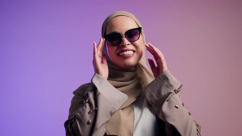 Enjoying modern accessories, gestures of femininity and beauty, attention to one's appearance and elegance, photography of a veiled Saudi Arabian Gulf woman trying out new sunglasses, an Emirati woman wearing an elegant abaya and contemporary black sunglasses, a colorful background.