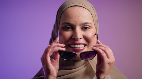 Concern for appearance and elegance. Photograph of a veiled Saudi Arabian Gulf woman trying out new sunglasses. An Emirati woman wearing an elegant abaya and contemporary black sunglasses. Enjoying modern accessories. Feminine and beautiful gestures. Colorful background.