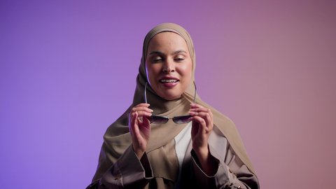 تصوير لإمرأة عربية خليجية سعودية محجبة تقوم بتجربة النظارات الشمسية الجديدة,  سيدة اماراتية ترتدي عباءة أنيقة ونظارة سوداء معاصرة, مفهوم الموضة والأناقة, الاستمتاع بإمتلاك اكسسوارات عصرية, إيماءات الانوثة والجمال, خلفية ملونة