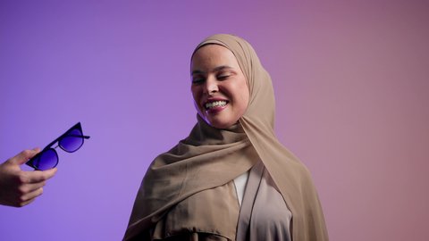 مفهوم الموضة والأناقة, الاستمتاع بإمتلاك اكسسوارات عصرية, تصوير لإمرأة عربية خليجية سعودية محجبة تقوم بتجربة النظارات الشمسية الجديدة, إيماءات الانوثة والجمال, سيدة اماراتية ترتدي عباءة أنيقة ونظارة سوداء معاصرة, خلفية ملونة