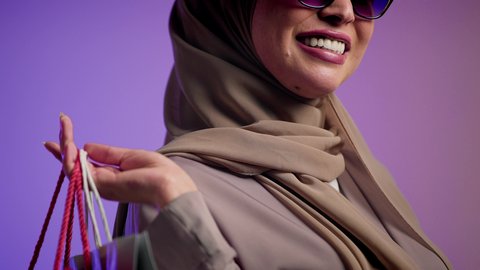 سيدة اماراتية تستفيد من العروض الموسمية والتخفيضات، الأثر الإيجابي للتسوق على السيدات، شراء المستلزمات والتبضع من المولات التجارية،  تصوير لسيده عربية خليجية سعودية مبتسمة ترتدي العباءه والحجاب ونظارة سوداء تحمل بيدها أكياس التسوق الملونه، خلفية ملونه