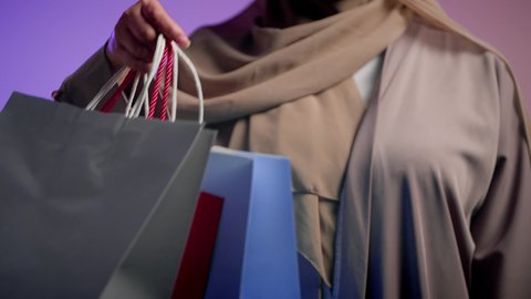 شراء المستلزمات والتبضع من المولات التجارية، سيدة اماراتية تستفيد من العروض الموسمية والتخفيضات، تصوير لسيده عربية خليجية سعودية مبتسمة ترتدي العباءه والحجاب تحمل بيدها أكياس التسوق الملونه، الأثر الإيجابي للتسوق على السيدات، خلفية ملونه