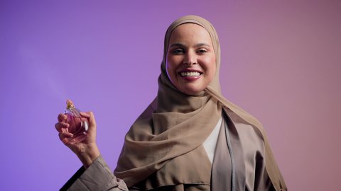 تصوير مقرب لامرأة عربيه سعودية خليجية محجبة مبتسمة تمسك بيدها زجاجة وتقوم برش العطر، سيدة اماراتية تستخدم المستحضرات النسائية, التعطر والتطيب بالعود والزيوت العطرية, التسويق لمنتجات العطور، خلفية ملونة