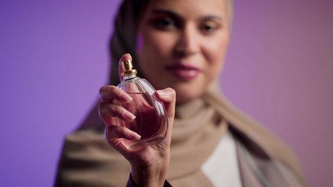 التعطر والتطيب بالعود والزيوت العطرية, التسويق لمنتجات العطور ، سيدة اماراتية تستخدم المستحضرات النسائية, تصوير مقرب لامرأة عربيه سعودية خليجية محجبة مبتسمة تمسك بيدها زجاجة وتقوم برش العطر، خلفية ملونة