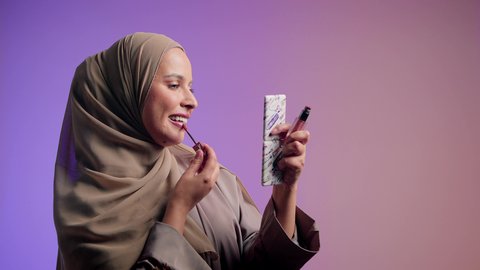 مفهوم الانوثه والاهتمام بالمظهر الخارجي،  التسويق لمنتجات التجميل ،سيدة اماراتية تستخدم أدوات تجميل نسائيه عصريه، مكياج وميكاب تتوريال، تصوير لفتاه عربيه سعودية خليجيه ترتدي العباءة والحجاب تقوم بوضع احمر الشفاة ، خلفيه ملونه