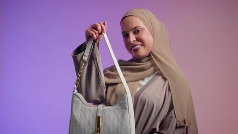مفهوم الموضة والأناقة، تصوير مقرب لشابة عربية سعودية خليجية محجبة تحمل بيدها حقيبة بإيماءات السعادة، عرض موديلات حقائب عالمية فاخرة، أزياء واكسسوارات نسائيه، سيدة اماراتية ترتدي العباءة الخليجية العصرية