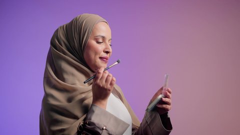 Makeup tutorial, the concept of femininity and attention to one's outward appearance, marketing for beauty products, a photograph of a Saudi Arabian Gulf woman wearing an abaya and hijab holding a blush brush, an Emirati woman using modern women's cosmetics, an upscale women's salon, a colorful background.